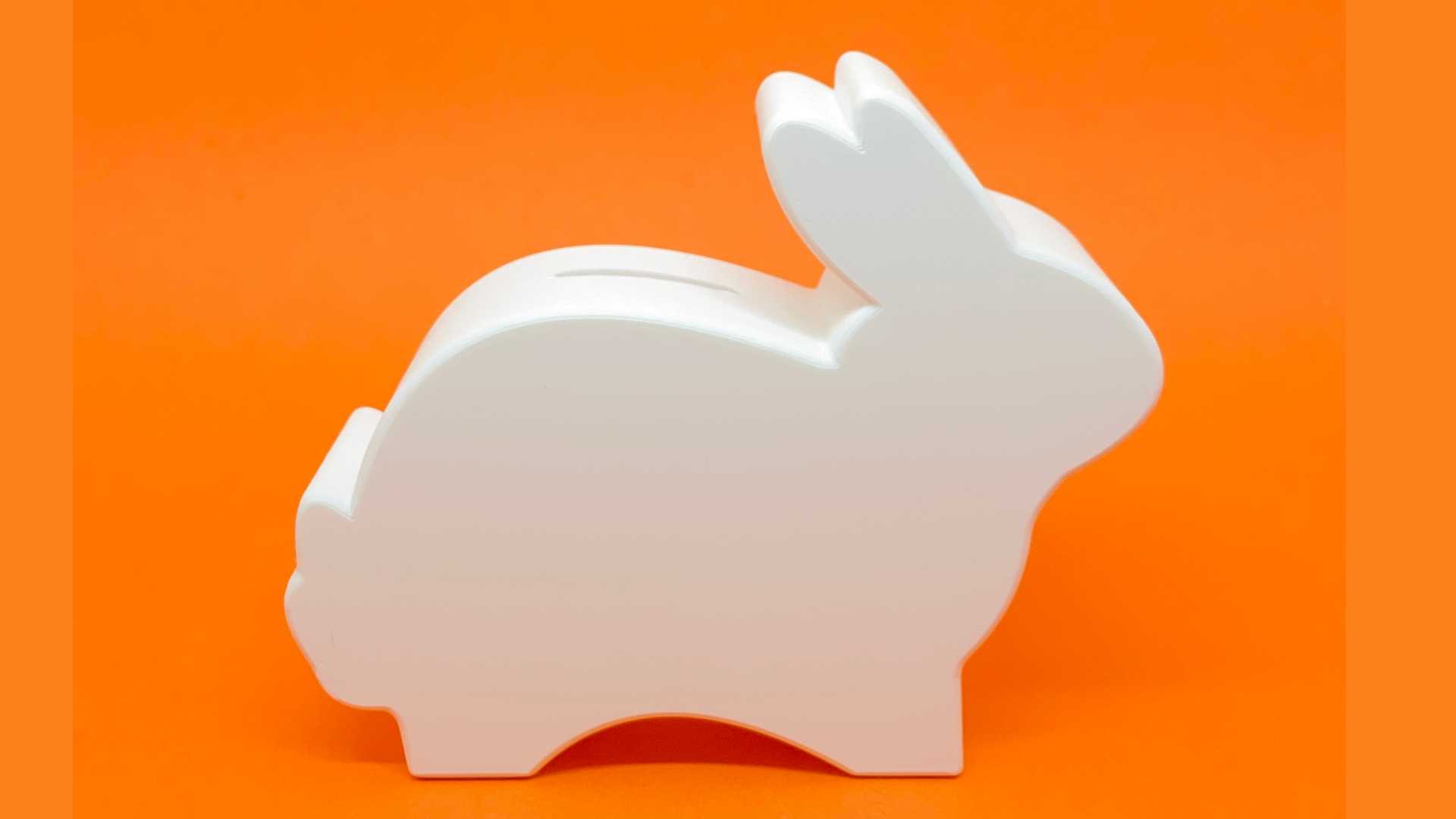 Fotografia de perfil de um cofre infantil em formato de coelho branco minimalista, posicionado sobre um fundo laranja vibrante. O coelho tem design clean e moderno, com bordas arredondadas, feito em impressão 3D sustentável.